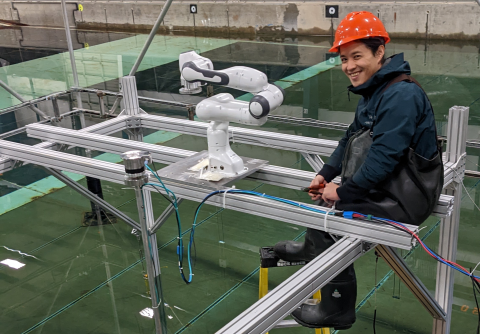 A student wearing a hard hat and waterproof attire sits on a structural beam above a tank of water. A robotic arm is attached to the beam next to him and a surfboard and offshore wind turbine tower float in the water below.