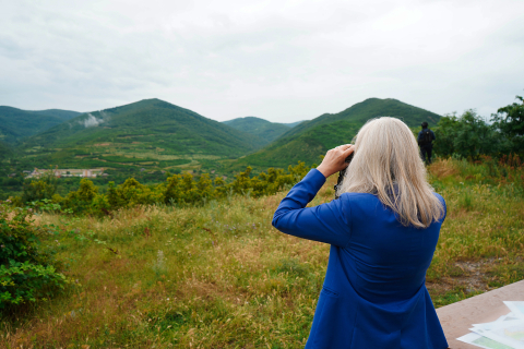 Shown from the back, NNSA Administrator Hruby looks over a hilly green landscape with a pair of binoculars.
