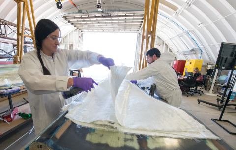 Two people in safety attire adjust fabric layers in a manufacturing facility