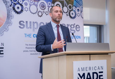 A man wearing a suit speaks from behind a podioum with a sign that says, “American Made.”