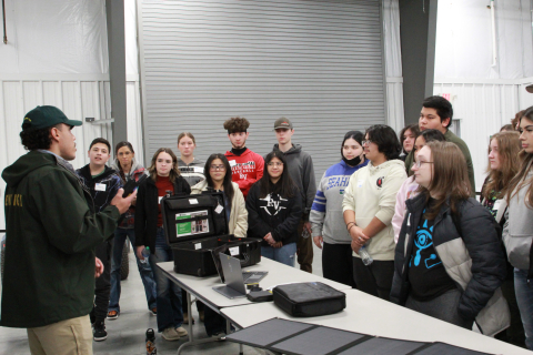 A group of students stand around a table and listen to a man talk 