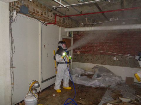 A worker wearing a hazmat suit in a stripped room sprays asbestos treatment throughout the room