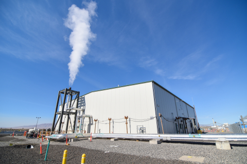 Large white facility building with a tall skinny chimney expelling smoke
