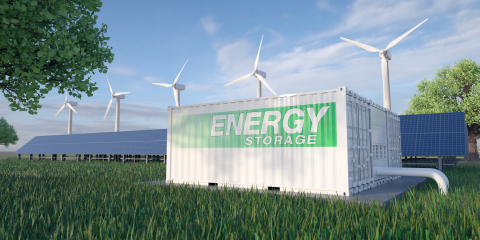 An energy storage container on the grass with solar panels and wind turbines in the background. 