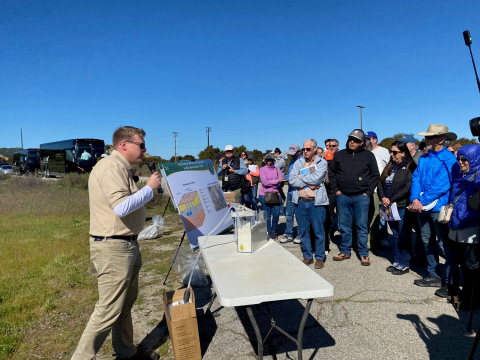 A man standing at a table with posters and science related models speaking to a crowd 