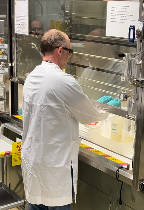 A man in a white lab coat and goggles opens a fume hood inside a lab and works inside of it