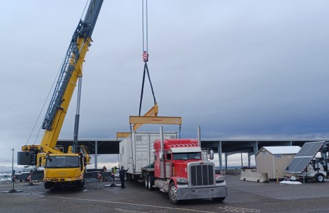 A crane unloading large boxes from the back of a hauler truck 