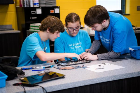 Three high school students sit and stand around a table and work on repairing a laptop
