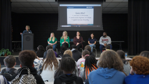 5 people sit in chairs on a stage with 1 standing beside them behind a podium. A PowerPoint is displayed behind them and a crowd watches and listens to them