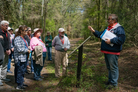 A group of people on a hiking trail in the woods stand around a read a plaque while a man stands behind it and talks to the group 