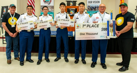 A group of students pose for a picture while holding prize checks and a trophy