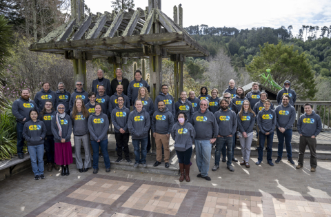 A group of more than 30 people of various genders and races, almost all wearing matching sweatshirts that have the Kbase logo on them. They are standing in a stone plaza with a small wooden structure behind them and further back, a sculpture of a preying mantis on a roof.