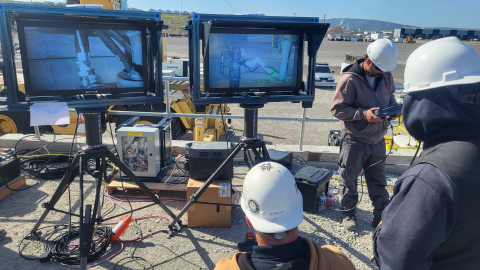 Two large screens displaying a machine. 3 workers with white construction hats stand around the screens 