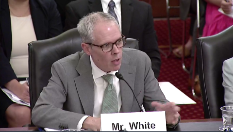 A man in a grey suit and glasses sits at a table with a name tag in front of him that reads "Mr. White"