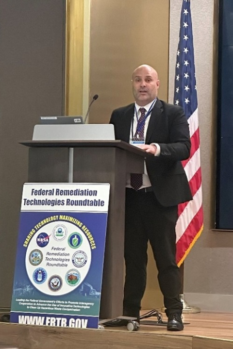A man stands behind a podium and speaks, an American flag hangs behind him