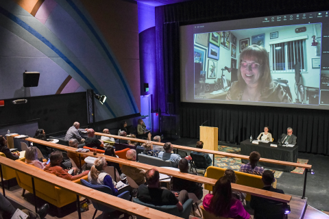 A view from behind of a filled auditorium with participants sitting at a panel in the front with a woman speaking via video call on the screen behind them