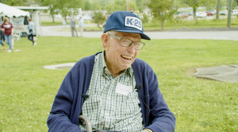 An old man sits on a chair with grass in the background behind him. The man is wearing a dark baseball hat