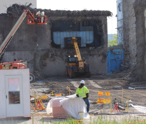Cranes working to demolish a building, an employee in a hardhat is seen in the foreground 