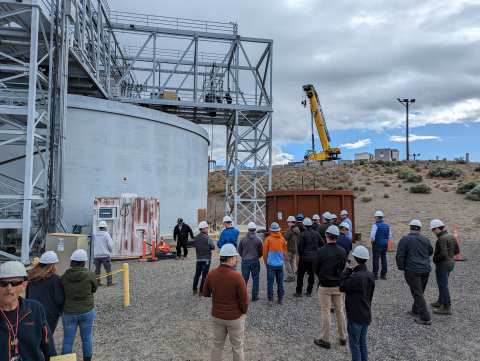 A large group of people in white hard hats look at circular facility building with metal ladders and construction vehicles around it