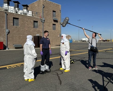 Two people in white hazmat suits and a man in firefighter boots and pants stand together, another person stands to the side and holds a large grey microphone over them 