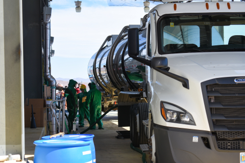 A large tractor-trailer truck getting fueled with gas by three men in green hazmat suits