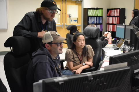 A group of people sit and stand around a computer and look at the computer screen