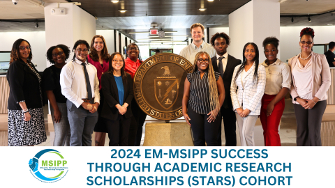 A group of young professionals pose for a group picture in front of the DOE seal in the DOE Headquarters building