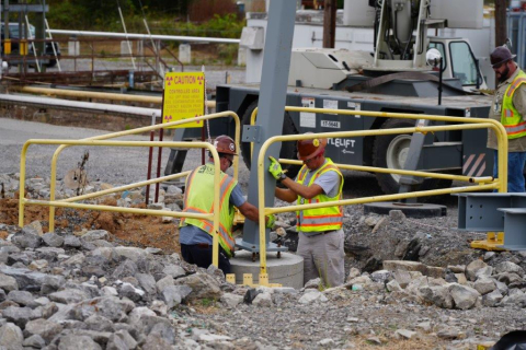 Two workers in safety vests and hard hats stand in a hole in the ground and install a steel beam