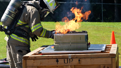 A firefighter in full gear demonstrates a fire with a metal object sitting on top of a large wooden box