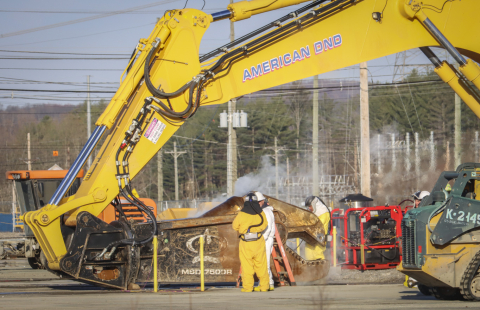 A large yellow construction crane vehicle works with employees standing around it in yellow suits