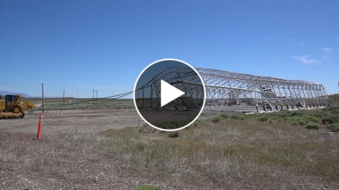 An open field with dried brown grass and a demolished building frame is in the background. A black and white play button is in the middle of the photo