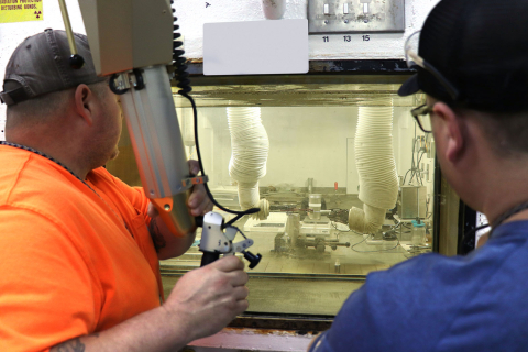 Two men face a machine behind glass, they control the robotic arm with a gray tool