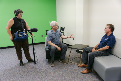 3 people in a room with one white wall and one green wall, 2 people sit and speak to eachother, another person operates a camera