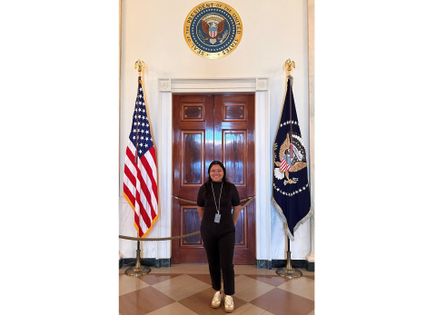 Cecilia Villegas standing in front of wooden door with American flag on one side and Flag of the President of the United States on the other side.