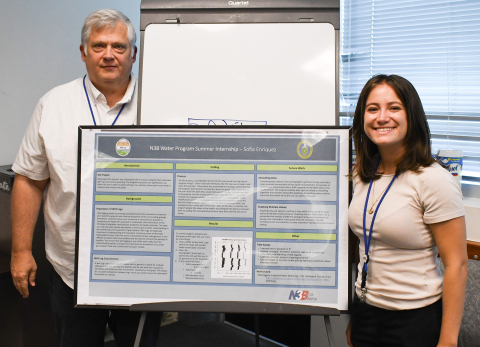 Two individuals stand beside a poster board and smile 