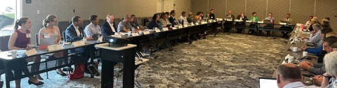 A large horseshoe shaped table in a room with people sitting in each chair and talking to each other