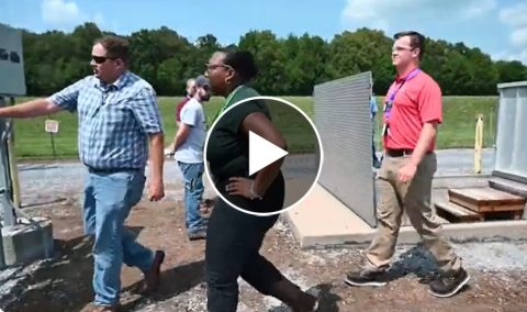 Individuals walk outside through a facility area, a play button is displayed in the middle of the image