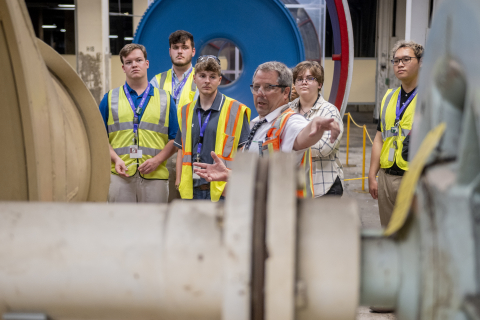 A group of student interns wearing yellow safety vests take a tour of a laboratory and listen to the employee speaking to them