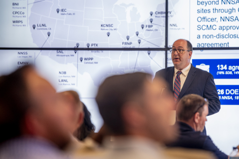 A man stands by a screen and gives a powerpoint presentation to the audience, who looks back at him