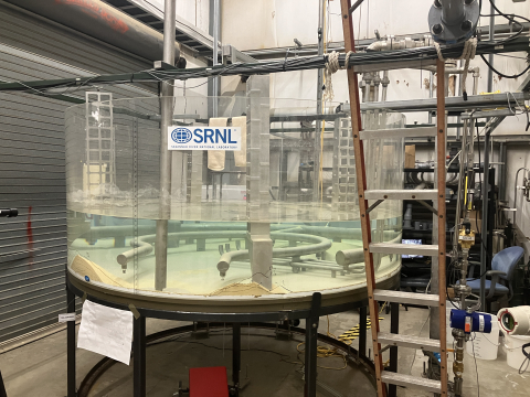 A large, round, glass table with clear liquid inside, it has pipes running through it and a ladder behind it 