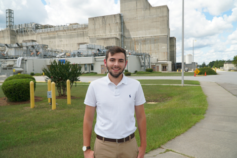 A man in a white shirt smiles and stands outside n front of a facility building