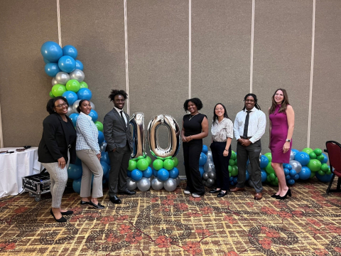 A group of interns stand and smile beside two silver balloons of the number 20 