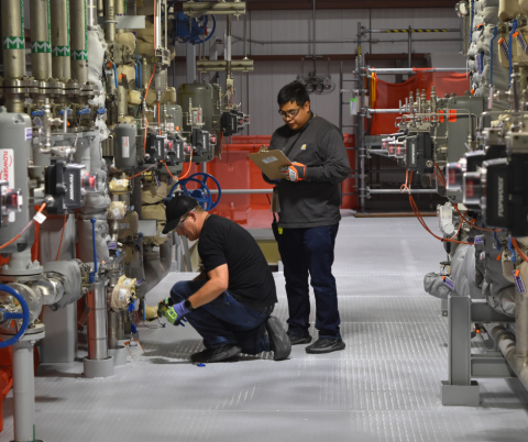 Two men inside a room with lots of pipes and pumps covering the wall, one man is crouched down looking at a pump and the other is holding a clipboard and writing