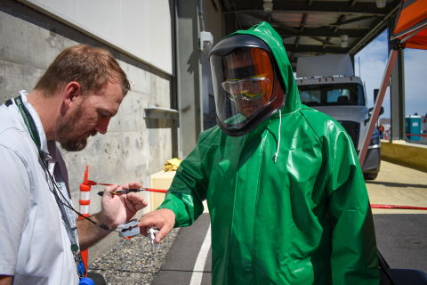 A man in a green hazmat suit holds his finger out and gets his heart rate measure by another man using a finger heart rate monitor clamp 