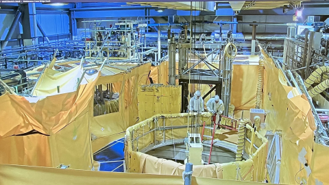 An aerial view of a factory building with two workers in hazmat suits performing work in the middle, the room is covered in yellow tarps