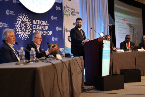 Department of Energy leaders sit at tables on two sides of a stage with another standing behind a podium