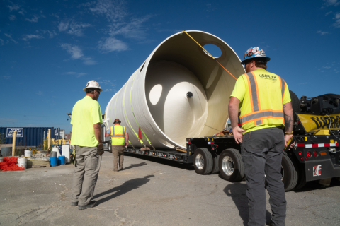 Employees in hard hats and yellow safety vests stand beside a large white tank being hauled on the back of a truck