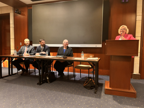 A group of DOE leaders sit at a table in the front of a room, a woman stands at a podium beside the table, they all smile at each other and the audience 