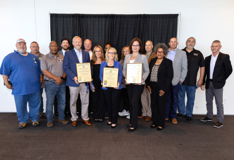 A group of people pose for a group photo, three individuals hold framed certificates 