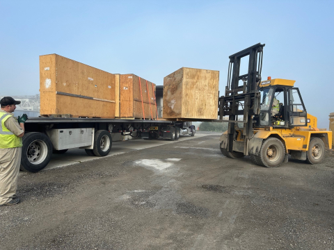 Workers operate a lift machine to move large wooden boxes onto a truck